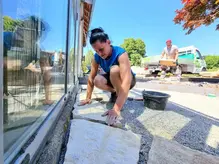 Gärtnerin verlegt Pflastersteine vor einem Gebäude unter blauem Himmel.