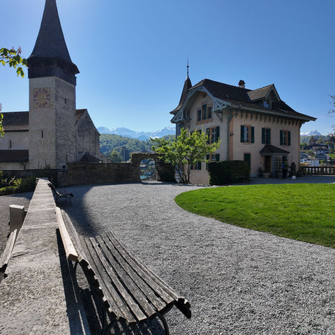 Church tower and building by a gravel path with mountains.