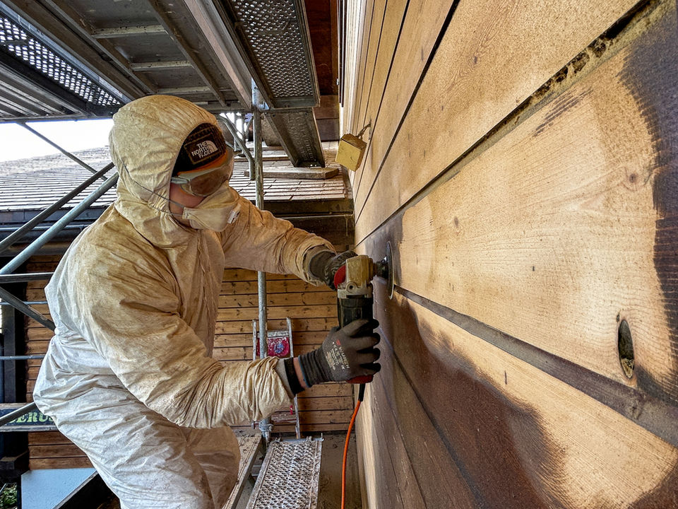 Handwerker im Schutzanzug schleift eine Holzfassade ab.
