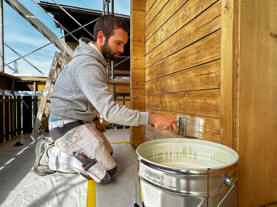Maler trägt mit Pinsel Lasur auf eine Holzfassade auf.