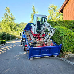Blauer LKW transportiert Minibagger auf einer Straße vor Hecke und Gebäude.