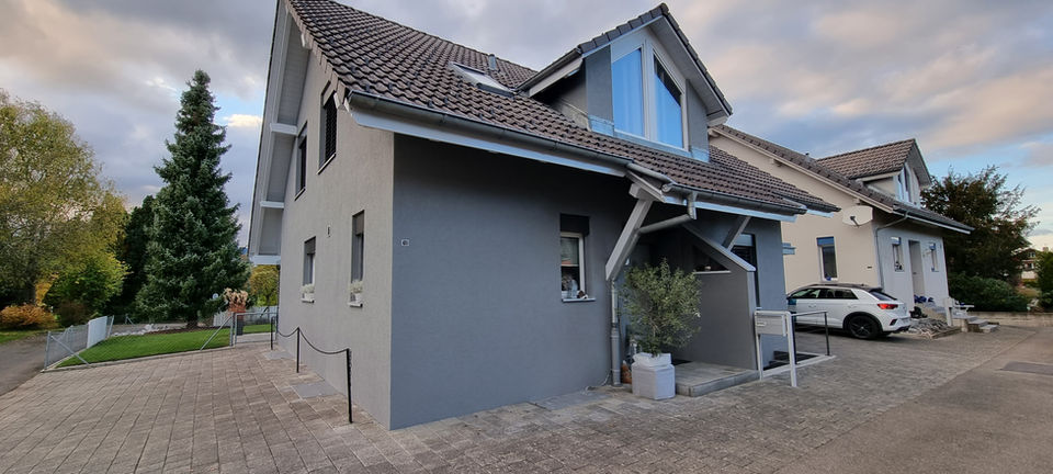 Modernes Einfamilienhaus mit grauer Fassade und Satteldach, davor gepflasterter Vorplatz und Pflanzen.