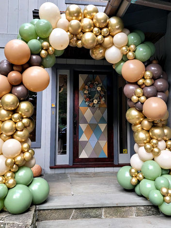 a very classy organic style balloon arch in front of a doorway to a home. The arch features a classic arch shape with different sized balloons. The colors shown are muted brown, cream, and green tones and brilliant gold balloons.