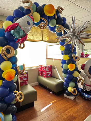 A balloon arch with an outer space theme, featuring dark blue balloons and accents of green and pale yellow. There are gold curly balloons, planet balloons, a large star, a rocket ship foil balloon and a 4' astronaut balloon