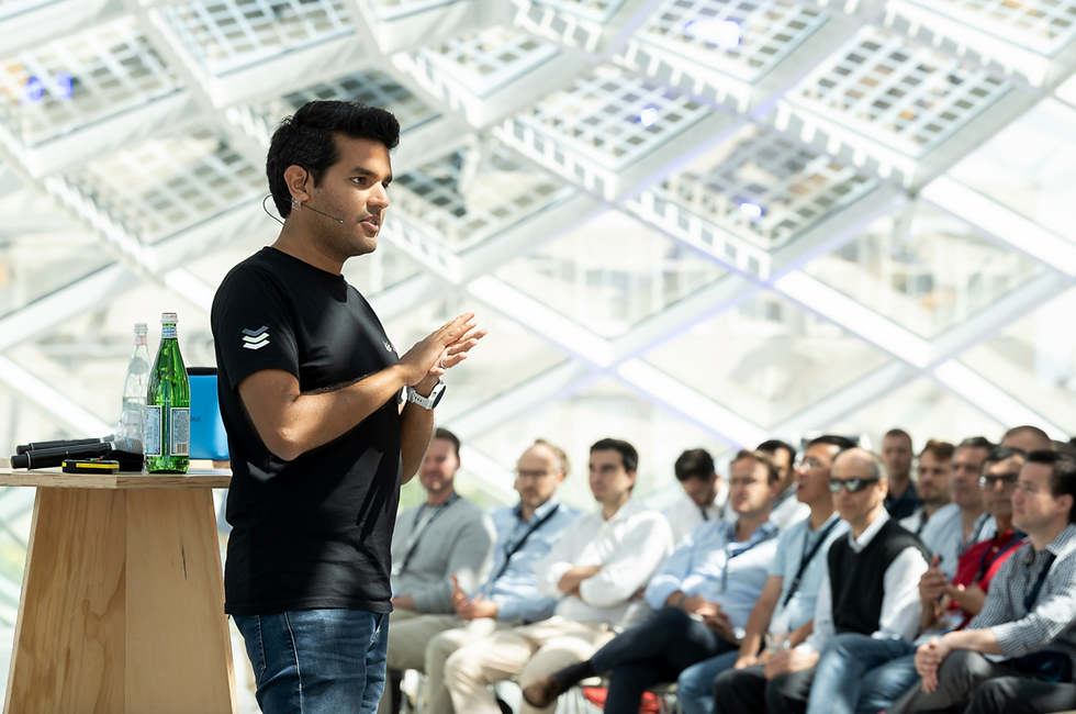 Man in black shirt and headset speaks to seated audience at a bright modern venue, gesturing beside a small table with bottles and mic - captured by professional photographer