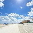 Sunny beach with blue umbrellas, a yellow lifeguard tower, and distant beachfront buildings under a wide sky of bright clouds
