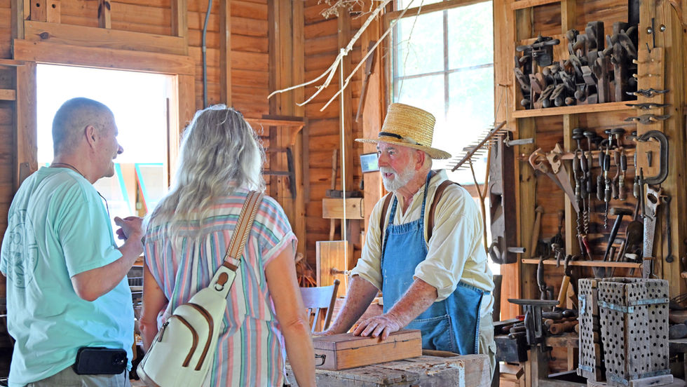 Older man in straw hat and blue apron talks to two visitors in a wooden workshop lined with hand tools.