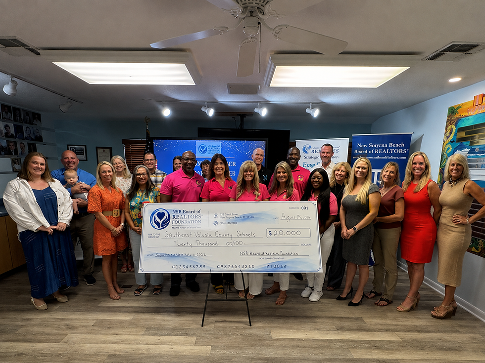 Real estate group poses indoors with a $20,000 check for Southeast Volusia County Schools, smiling by NSB Board of REALTORS banners.