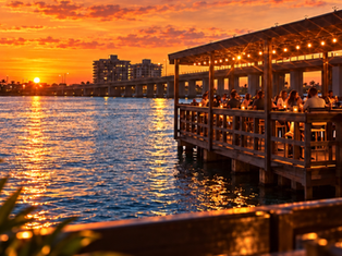 Sunset over a waterfront restaurant patio with diners, glowing lights, rippling water, and orange clouds over the bay