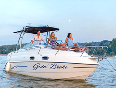 Three adults relax on a white yacht named Goin’ Broke on calm water at dusk, with a wooded shoreline in back.