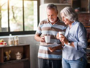 Grey-haired couple smiling, holding mugs and reading a newspaper in a cozy kitchen with large windows and soft natural light.