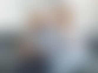 An older man in a blue shirt sits on a grey couch, looking pensive. A woman beside him, in a white blouse, rests her hand on his shoulder, appearing supportive but worried.