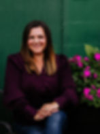 Smiling woman in a purple blouse sits with hands folded, next to vibrant pink flowers against a green wall. Relaxed and cheerful mood.