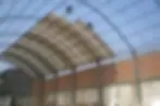 Fase avanzada de techado sobre un sistema de arcos parabólicos reticulados de gran luz apoyados en columnas de concreto. El proceso muestra el despliegue e instalación consecutiva de planchas de cobertura aligerada, fijadas con precisión sobre las correas superiores, estructurando una superficie curva hermética y aerodinámica que maximiza el volumen interior útil del almacén.