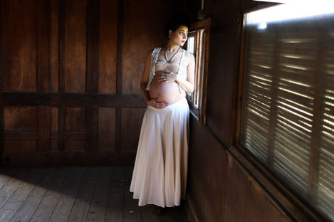 אישה הרה עומדת ליד חלון, מחזיקה בבטנה בתוך חדר עץ, צילומי הריון.