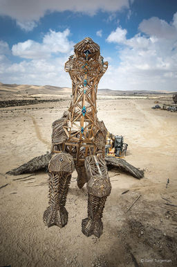 פסל אמנותי גדול במדבר, מבנה ייחודי, רקע שמיים בהירים, עם פרטים בולטים בשטח.