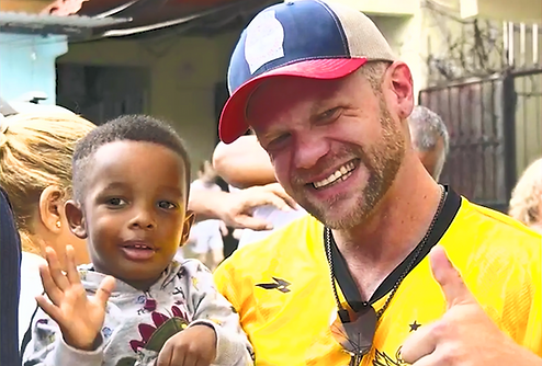 Michael Urbanowicz smiling and giving a thumbs up while holding hands with a young boy at the Panama Faith Over CP Kick-Off event. The boy is holding a drink and smiling, capturing a moment of connection and joy in the vibrant outdoor setting.