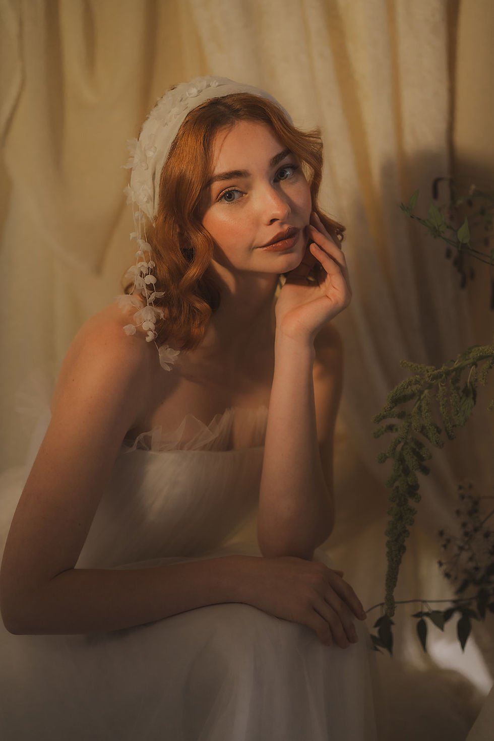Thumbnail: Young woman in white dress, wearing a delicate silk chiffon headband featuring delicate threads dangling down with 3D silk flowers, posing with hand on face.