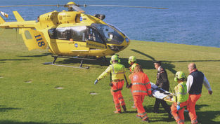 Rescuers in orange and green gear carry a stretcher to a yellow emergency helicopter beside a lake. The mood is urgent and focused. travel insurance, איתור וחילוץ על ידי מסוק 