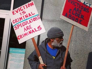 POOR Magazine member holding signs saying "Eviction is criminal / Speculators are criminals" and "Ellis Act evictions are elder abuse" outside the San Francisco Hall of Justice