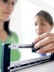 Close-up of a hand adjusting a scale. A person in a green shirt stands beside. Bright, clinical setting conveys focus and professionalism.