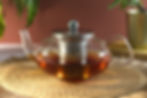 A glass teapot with a silver strainer with brewed tea in the teapot. Red background with plans and sitting on a brown mat.