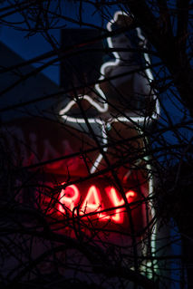 À travers des branches d'arbres nues, le néon de l'enseigne du bar Miss Villeray. Les seules parties allumées sont une silhouette de serveuse et les lettres R, A et Y.