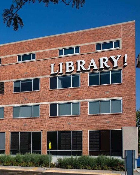 exterior of the Boise Public Library's main branch. It's a red brick building with three rows of horizontal windows and Library! in white serif font. The sky is clear and blue and trees and bushes are also visible.