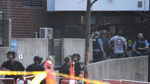 Police and tactical officers gather outside a brick building behind yellow caution tape and barricades.
