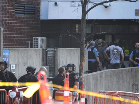 Police and tactical officers gather outside a brick building behind yellow caution tape and barricades.