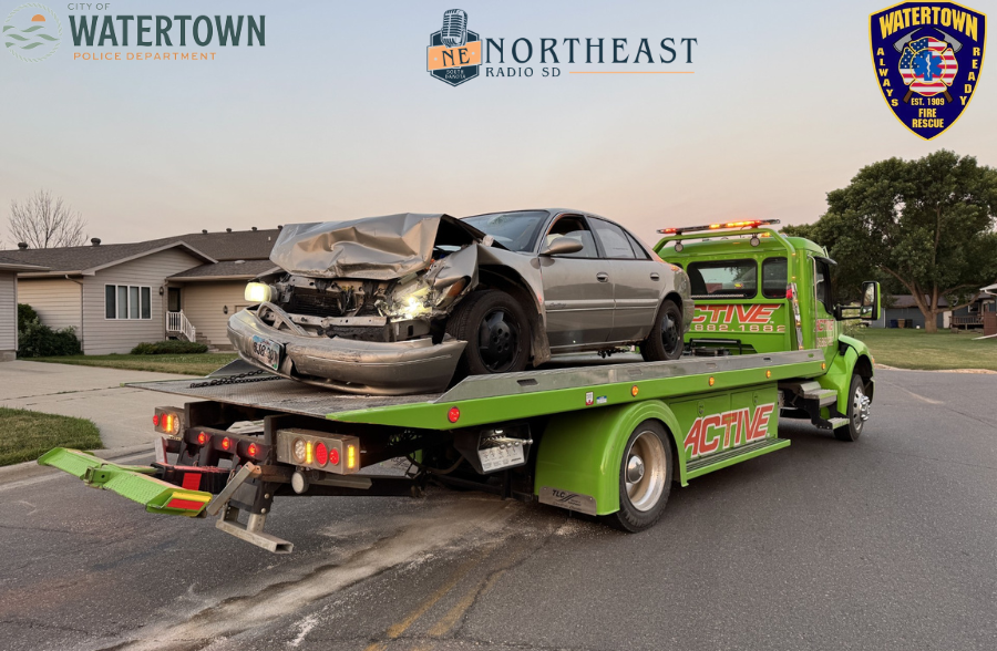 Damaged sedan loaded on bright green tow truck on a neighborhood street at dusk, with Watertown police and fire logos above.