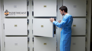 Man in a blue lab coat opens a vault door in a wall of white radio lockers labeled Northeast Radio SD.