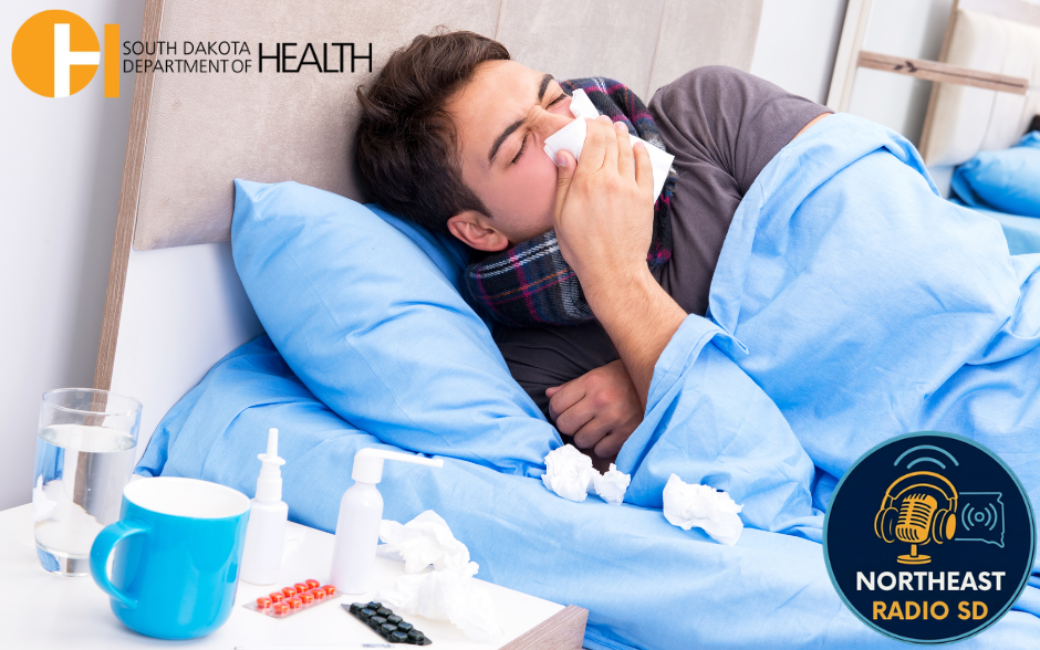 Man with cold in bed, blue blanket, using tissue. Meds and glass on table. South Dakota Dept. of Health and Northeast Radio SD logos visible.