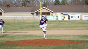 Baseball pitcher in blue uniform mid-windup on the mound, with teammate at left and sponsor signs on a sunny field.