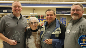 Four people smiling, one holding an award, indoors with banners and signs in the background. Casual attire, warm and celebratory mood.