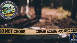 Crime scene with yellow tape reading "DO NOT CROSS." Blurred figures in the forested background. South Dakota seal and radio logo present.