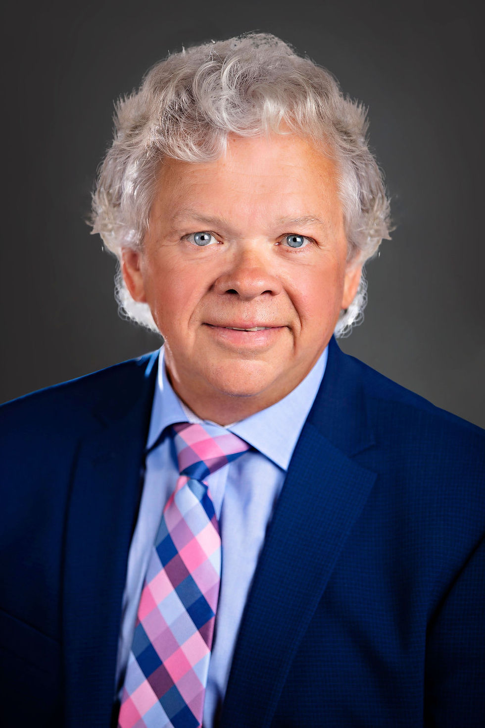 Portrait of a gray-haired man in a navy suit and pink-blue plaid tie, smiling against a dark gray studio background.