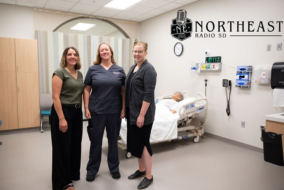 Three smiling women stand in a medical training room beside a hospital bed and mannequin; Northeast Radio SD logo on wall