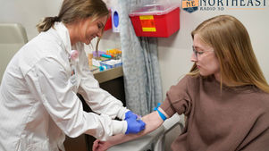 Nurse in white coat draws blood from a young woman in a clinic, with Northeast Radio SD logo in the corner.