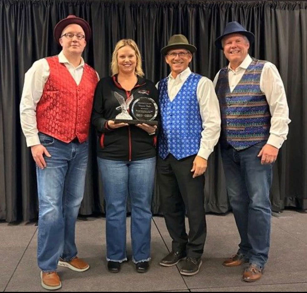 Four people pose onstage, three men in colorful vests and hats, one woman holding a trophy. Black curtain background, smiling faces.