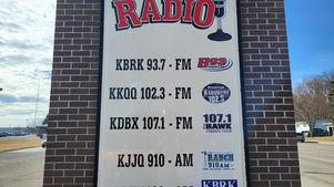 Signboard for Brookings Radio lists stations KBRK 93.7 FM, KKQQ 102.3 FM, KDBX 107.1 FM, KJJQ 910 AM, KBRK 1430 AM. Brick wall background.