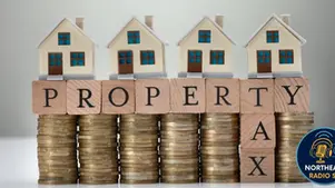 Toy houses on stacked coins spell "PROPERTY TAX" with wooden blocks. Logo in corner reads "NORTHEAST RADIO SD." Neutral background.