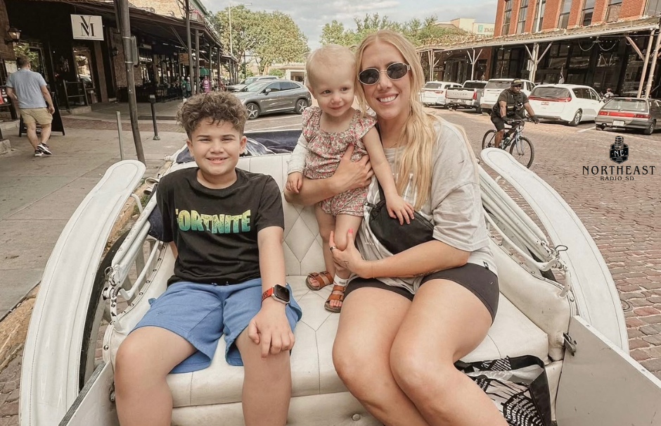 Smiling family in a white carriage on a brick street; boy wears a Fortnite shirt, woman holds toddler, downtown shops behind.