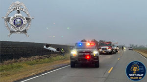 Police vehicles with flashing lights respond to an overturned white car in a field. Overcast sky. "NORTHEAST RADIO SD" logo visible.