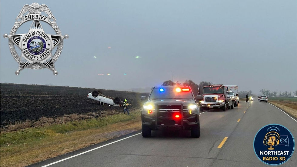 Police vehicles with flashing lights respond to an overturned white car in a field. Overcast sky. "NORTHEAST RADIO SD" logo visible.