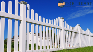 White picket fence against blue sky, houses in background. Grass in foreground. Northeast South Dakota Radio SD logo at top right.