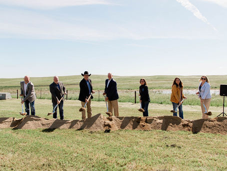 Six people hold shovels at a groundbreaking in a grassy field under a bright sky, looking formal and celebratory.