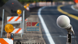 Roadwork barriers on a curving highway with a microphone foreground and SD Department of Transportation, Northeast Radio SD logos.