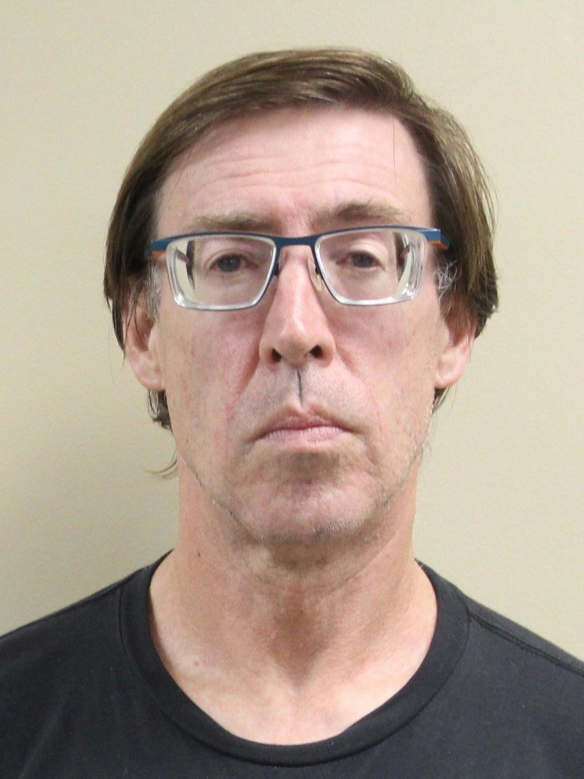 Mugshot-style portrait of a middle-aged man with glasses and a neutral expression against a beige background.