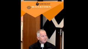Man in a Watertown 911 shirt speaks into a microphone during a Northeast Radio SD livestream, with orange promo banners and event text.